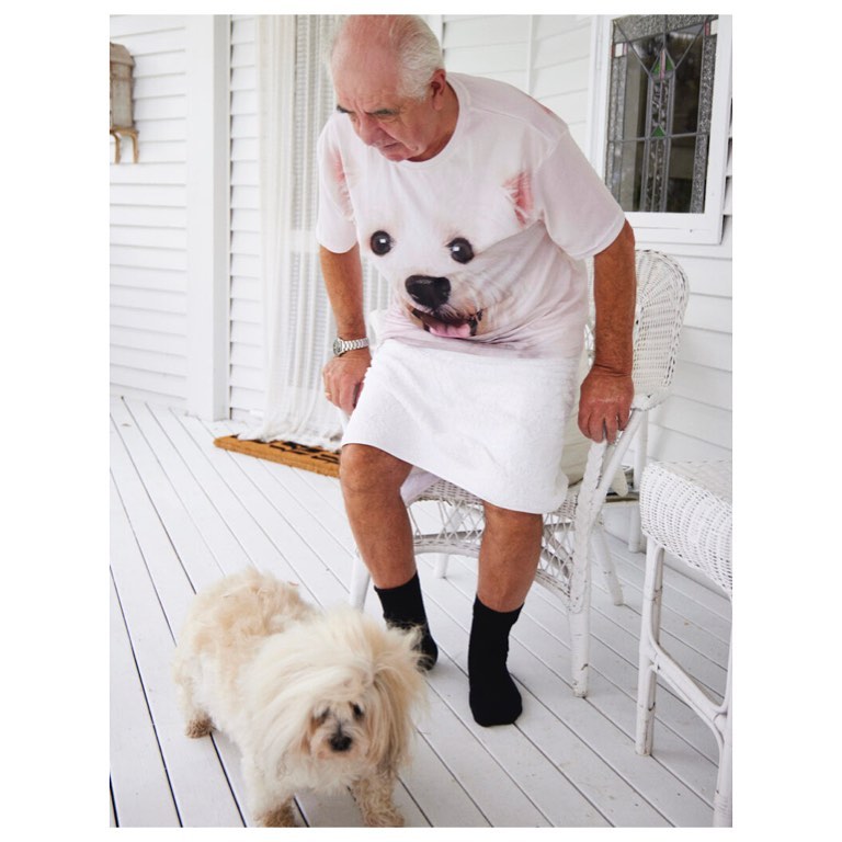 Elderly man in a dog-printed shirt stands on a porch next to a small white dog, creating a whimsical and lighthearted scene.