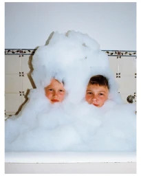 Two young boys partially submerged in a large mound of white soap suds inside a tiled bathtub, photographed by Joseph Molines.