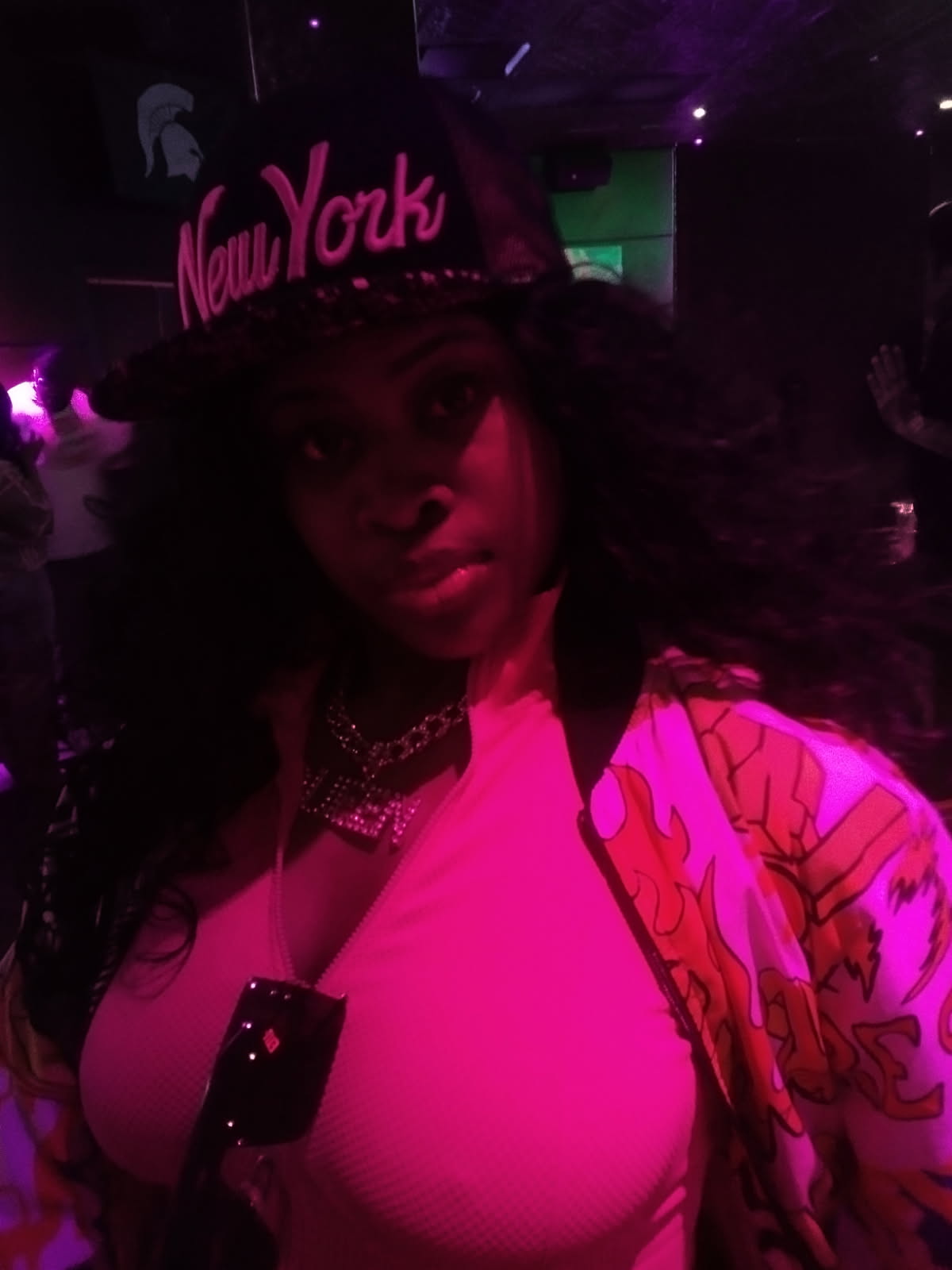Portrait of an adult woman with dark hair wearing a black cap embroidered with 'New York' at a nightclub with pink lighting.