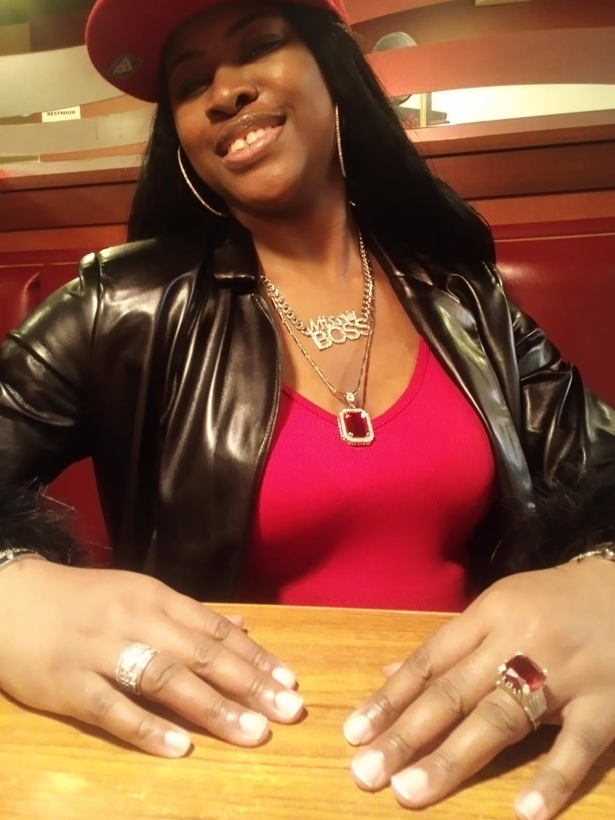 Smiling African-American woman in a red cap and black jacket at a table wearing multiple pieces of jewelry and rings inside.