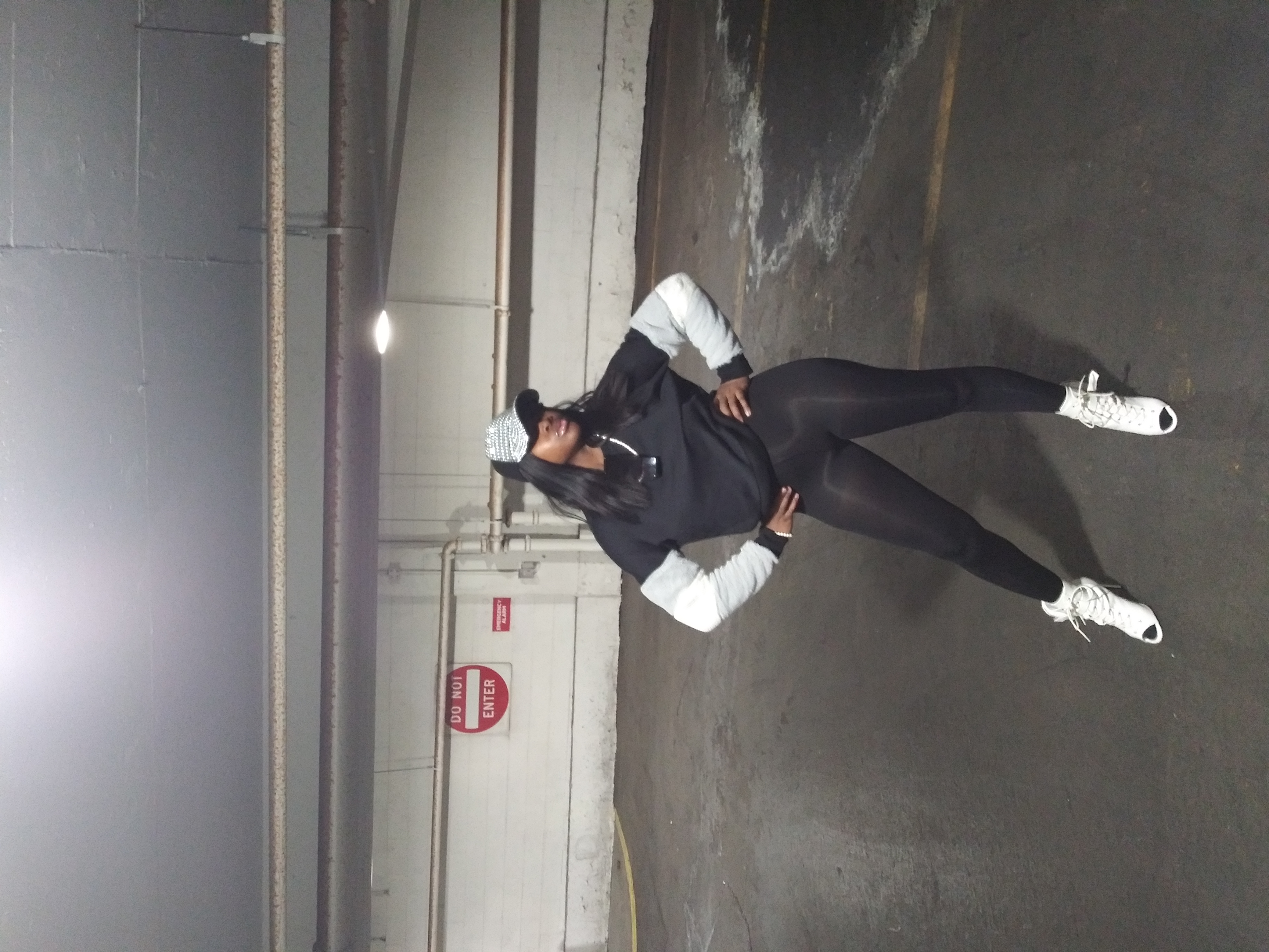 Woman in casual street style poses in a parking garage wearing black leggings, a sweatshirt with a hat and white sneakers.