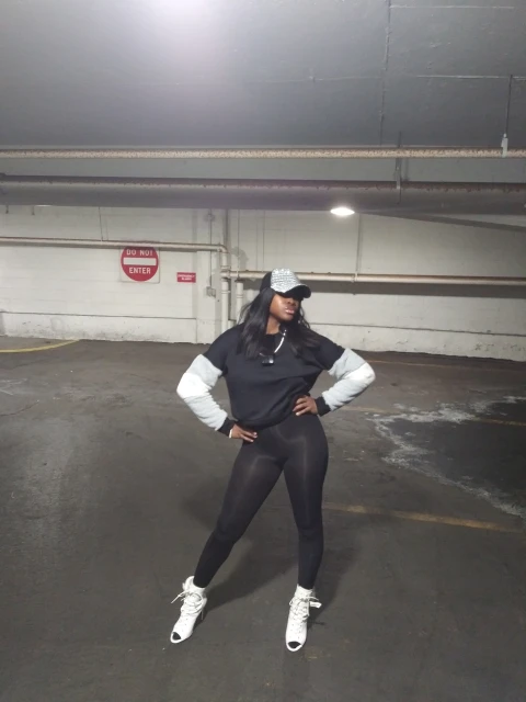 Woman in casual street style poses in a parking garage wearing black leggings, a sweatshirt with a hat and white sneakers.