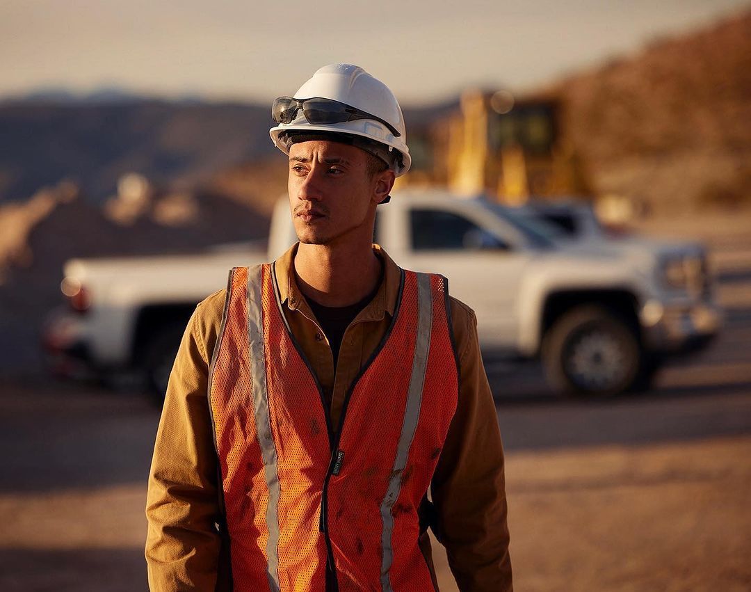 Man in safety vest and hard hat looks left on outdoor work site with truck and construction equipment in background in natural light.