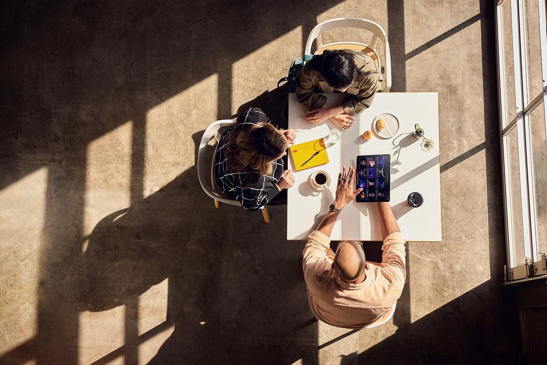 Overhead shot of three people at a white table using a tablet, with light and shadows creating an urban office setting.