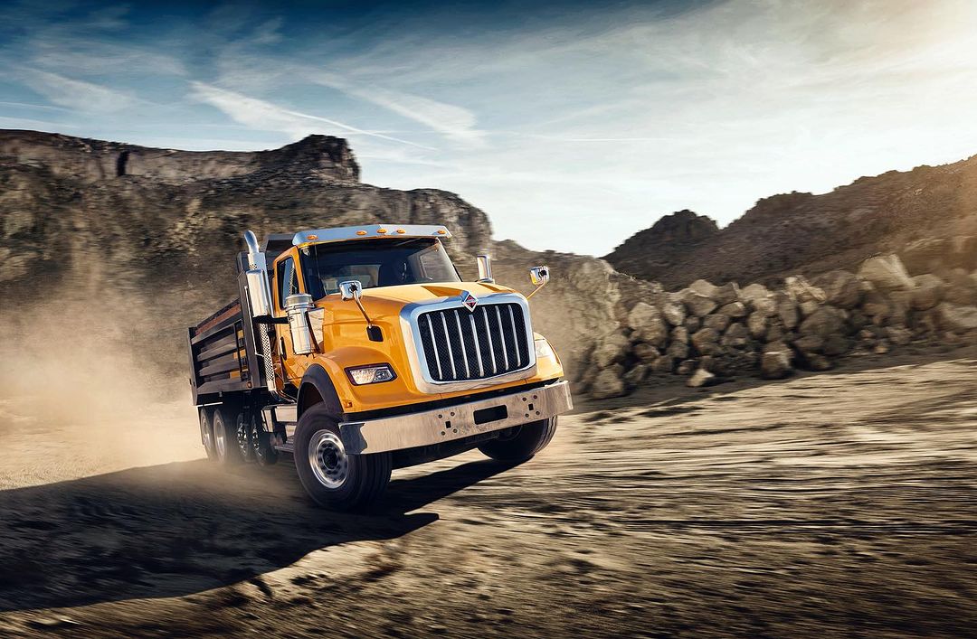 Yellow dump truck driving on a dirt road with a rocky landscape and blue sky in the background, capturing a rugged outdoor scene.