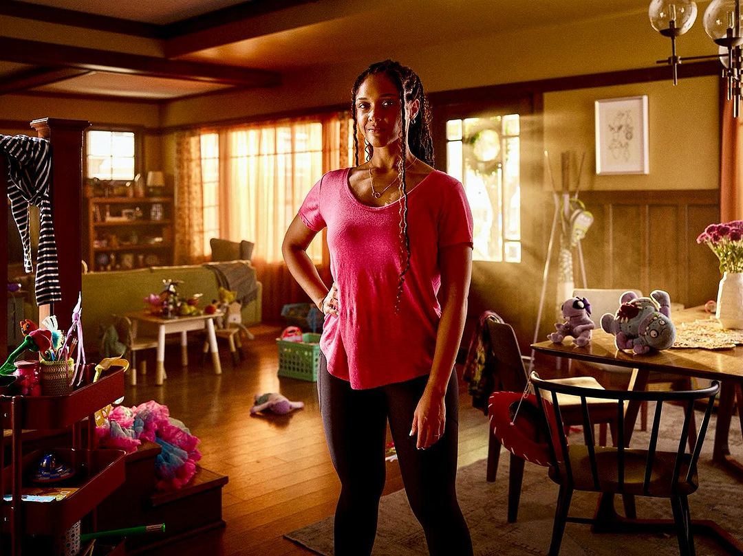 Woman in pink shirt stands confidently in a cluttered living room, showcasing a casual home setting with a blend of personal items.