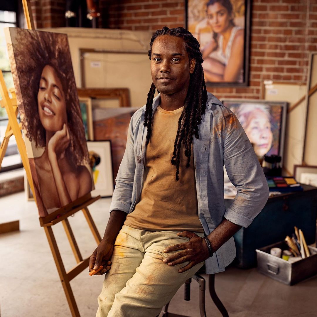An artist with dreadlocks sits next to his vibrant portrait of a smiling woman in his studio, surrounded by paintings and art supplies.