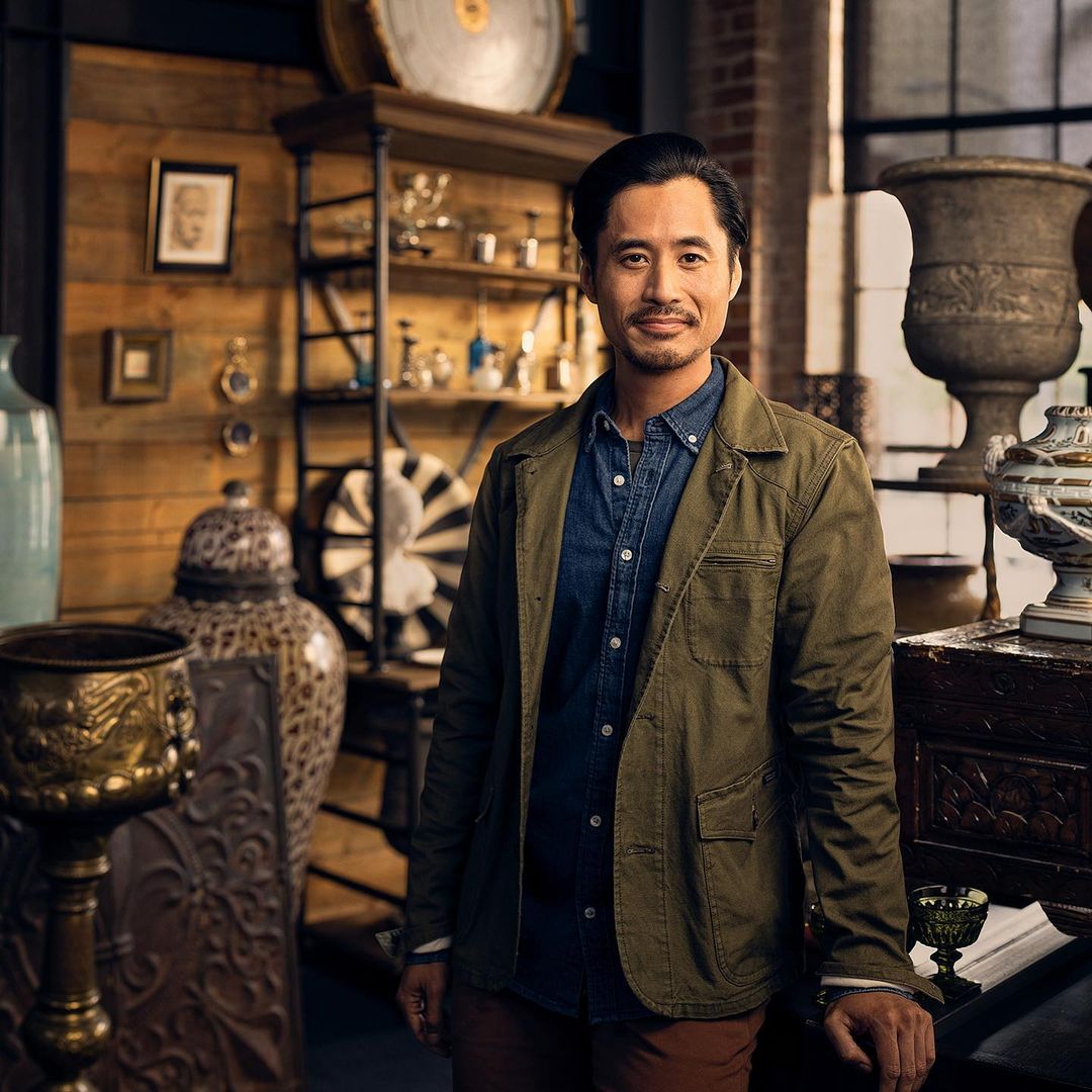 Portrait of a smiling adult Asian man in a green jacket, standing indoors surrounded by antique objects and decor in a store.