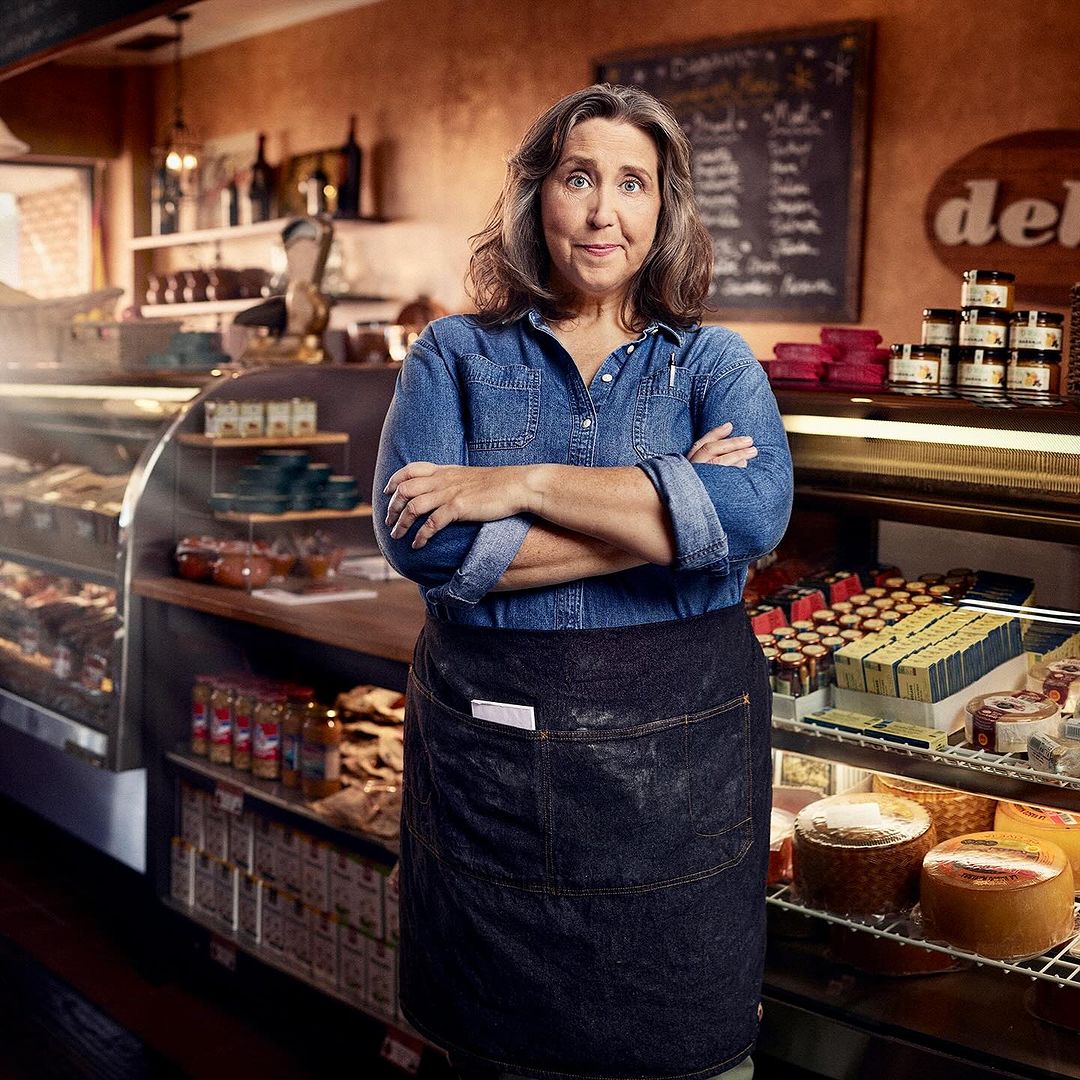 Mature woman in a blue denim shirt and black apron stands with arms crossed in a deli or specialty food shop, smiling confidently.