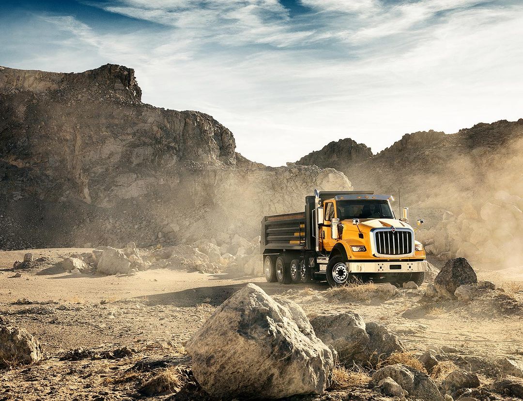 An orange International construction dump truck drives through a gravel pit with a quarry in the background under a sunny sky.