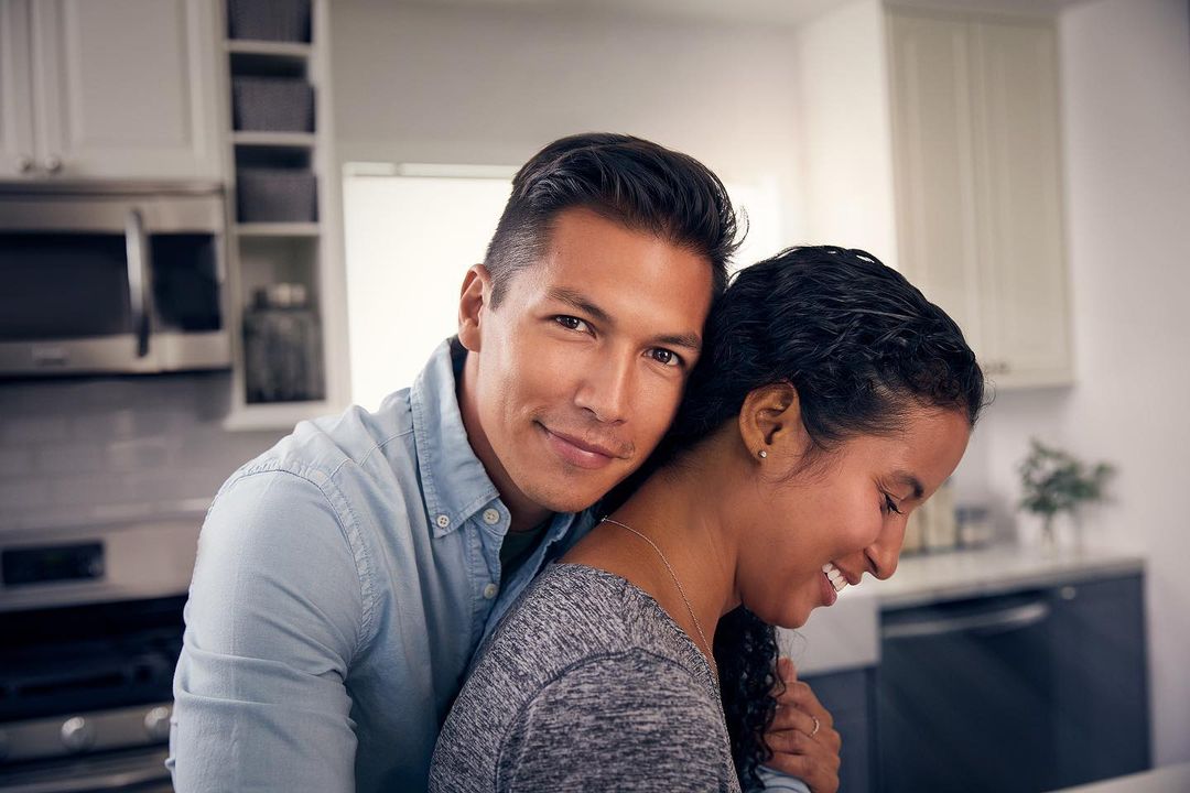 Smiling couple embraces in a brightly lit kitchen with white cabinets and stainless steel appliances, showcasing a modern home interior.