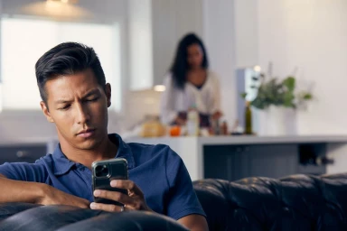 Man seated on a dark leather couch looking down at his smartphone in a domestic interior with a woman standing blurred in the background