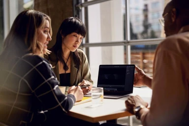 Three people collaborating around a laptop at a table indoors in New York by photographer Joseph Puhy