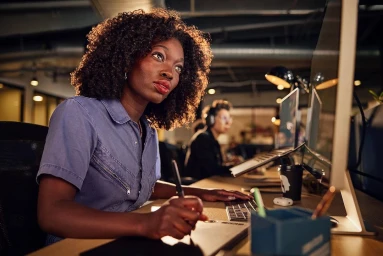 Black woman with curly hair seated at a desk writing on a tablet with a stylus in an office lit by warm lighting.