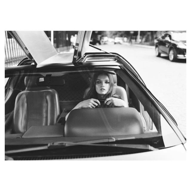 Monochrome portrait of a woman behind the wheel of a DeLorean car with gull-wing doors open on a city street, with other vehicles nearby.