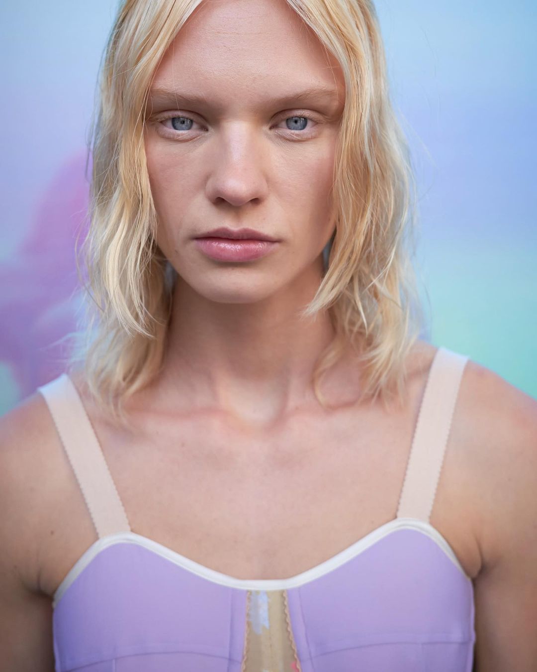 Close-up studio portrait of a fair-skinned adult woman with blonde hair, blue eyes, wearing a purple bra top and a neutral makeup look.