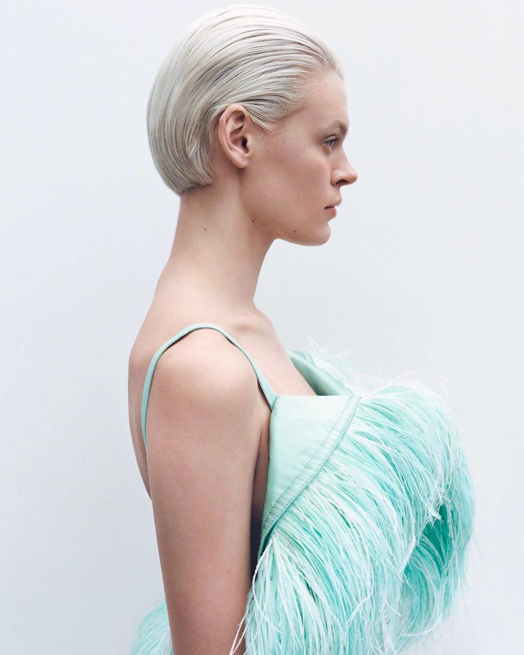 Profile of a woman with short platinum blonde hair in a light green feathered dress against a white background in a studio shot.