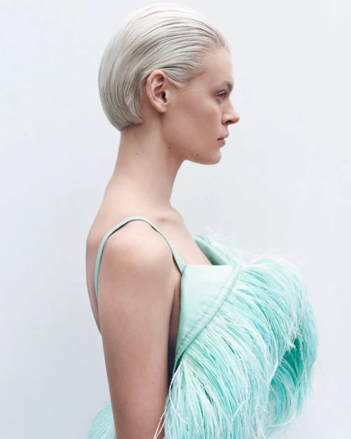 Profile of a woman with short platinum blonde hair in a light green feathered dress against a white background in a studio shot.