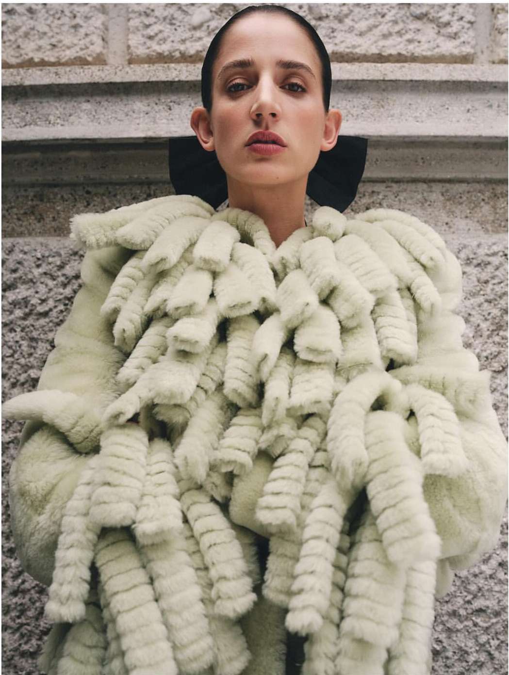 Woman wearing a light green, fur-like coat with a black collar standing indoors against a textured wall. Fashion style, medium shot.