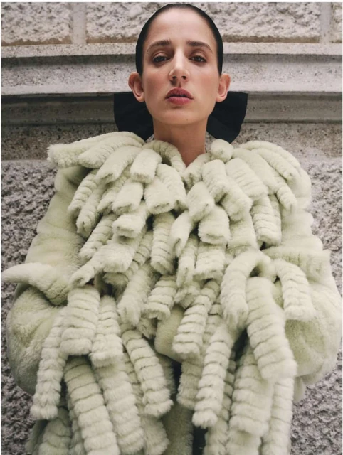 Woman wearing a light green, fur-like coat with a black collar standing indoors against a textured wall. Fashion style, medium shot.