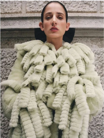 Woman wearing a light green, fur-like coat with a black collar standing indoors against a textured wall. Fashion style, medium shot.