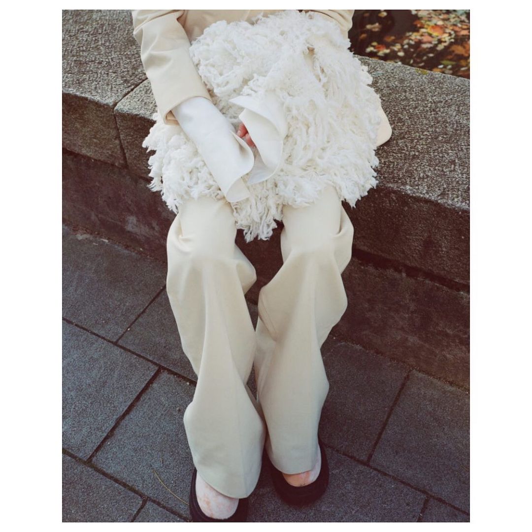 Fashionable person in cream-colored apparel sits on a stone ledge in an outdoor setting, showcasing chic style with black shoes.