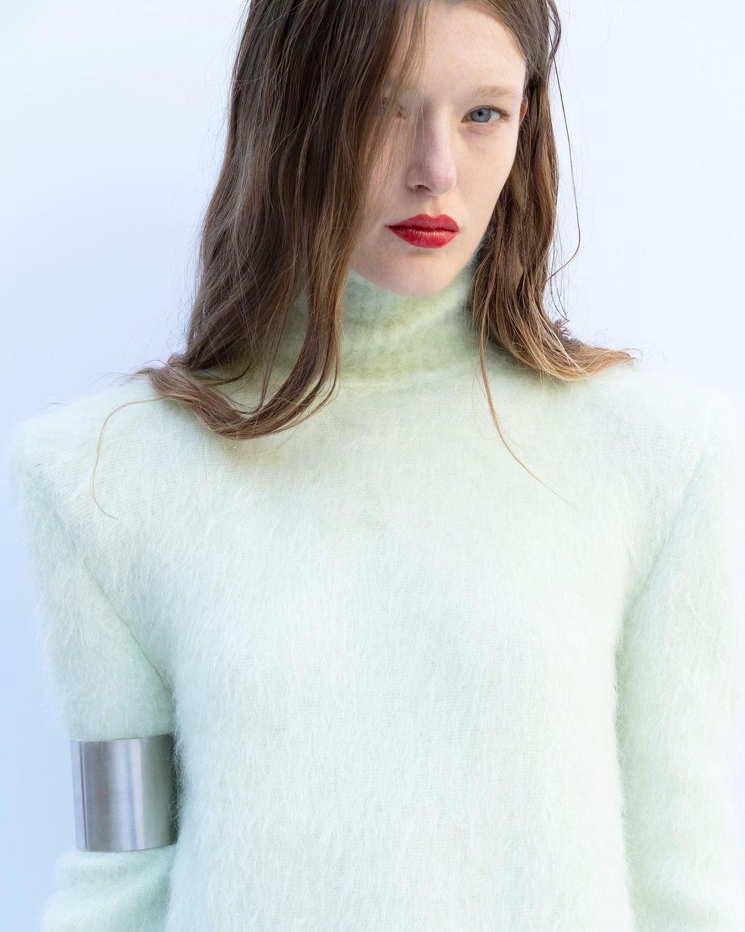 Close-up of a female model in a light green sweater with red lipstick and a metal arm cuff, posing against a light background.