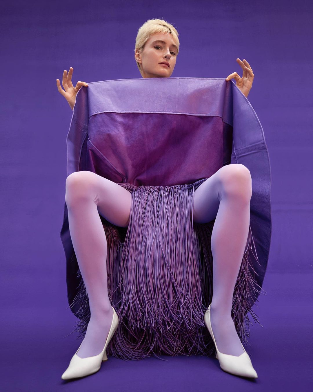Woman with short blonde hair sits on chair, posing with purple fabric, light purple tights, and white heels against purple backdrop.