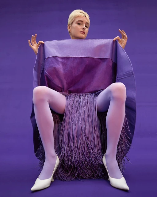 Woman with short blonde hair sits on chair, posing with purple fabric, light purple tights, and white heels against purple backdrop.