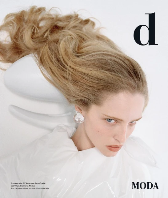 Fashion editorial shot: a female model with blonde hair poses in white with a magazine including the word 'Moda' visible.