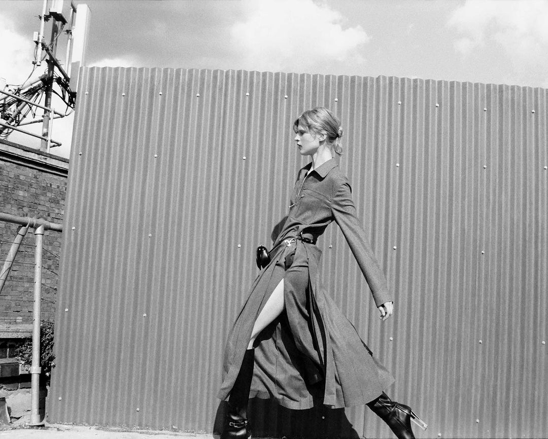 Monochrome fashion shot of a slender woman in a long gray coat walking outdoors next to a corrugated metal wall, wearing black heeled boots.