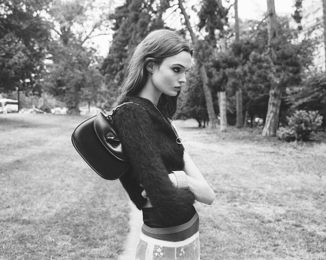 Black and white portrait of a woman in a dark fuzzy sweater standing in a park with a black shoulder bag slung behind her.