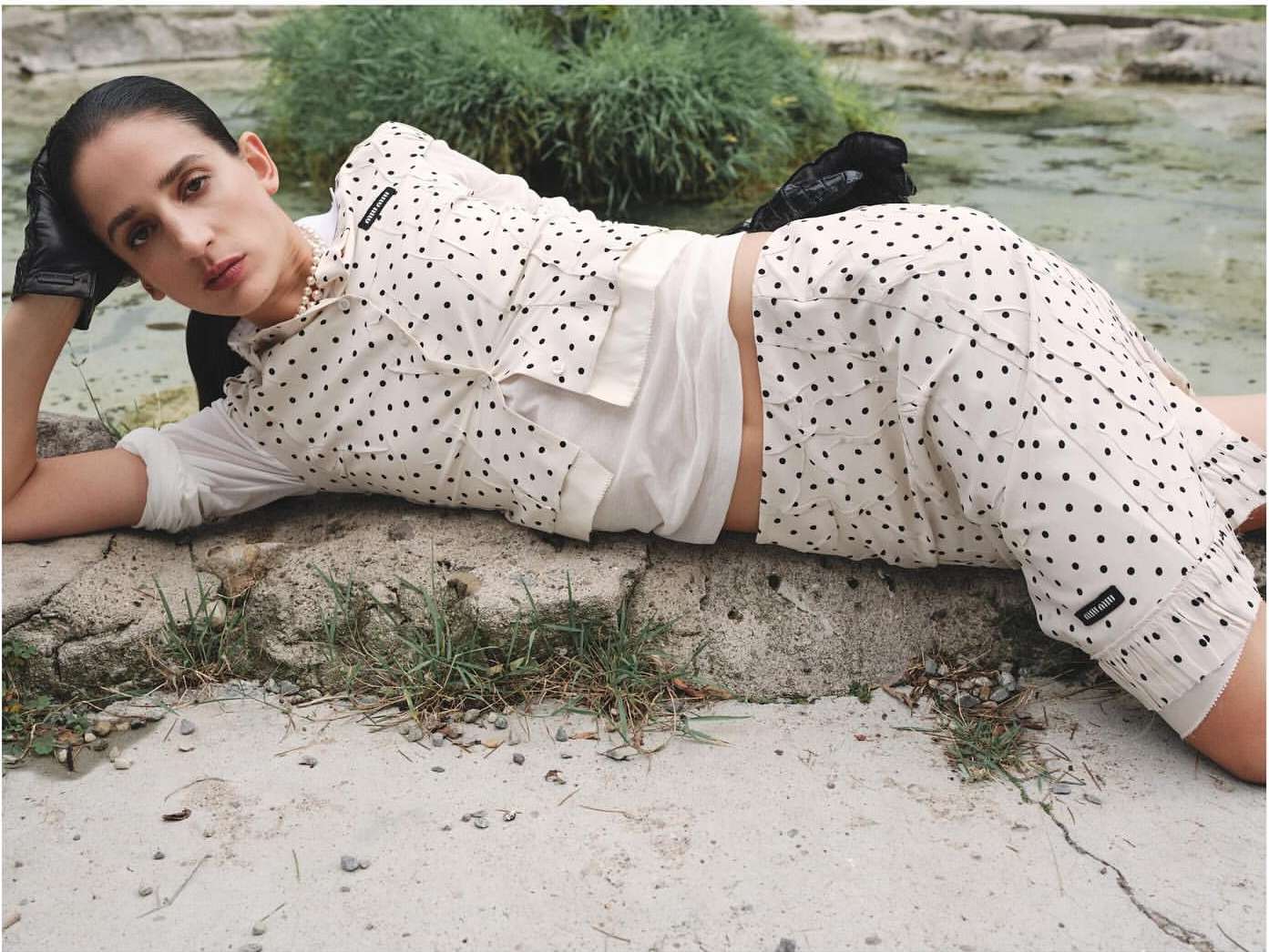 Adult woman with polka dot fashion lying on a stone surface with green plants. Editorial fashion style with a neutral color palette.