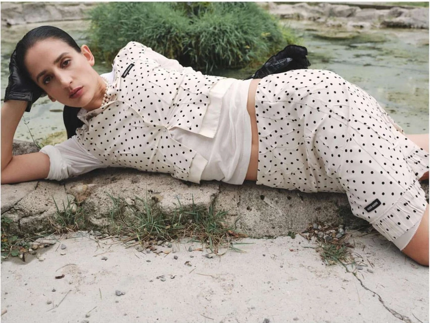 Adult woman with polka dot fashion lying on a stone surface with green plants. Editorial fashion style with a neutral color palette.