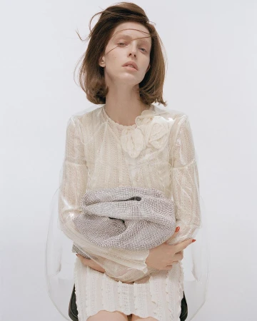 Model seated in transparent chair holding a silver mesh handbag wearing cream lace dress
