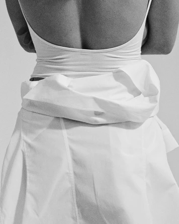 Monochrome rear view of a woman wearing a white tank top and wrapped white skirt in a minimalist studio setting.