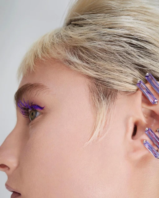 Close-up studio shot of a light-skinned woman with blonde hair, purple mascara, and lavender gem ear jewelry against a white backdrop.