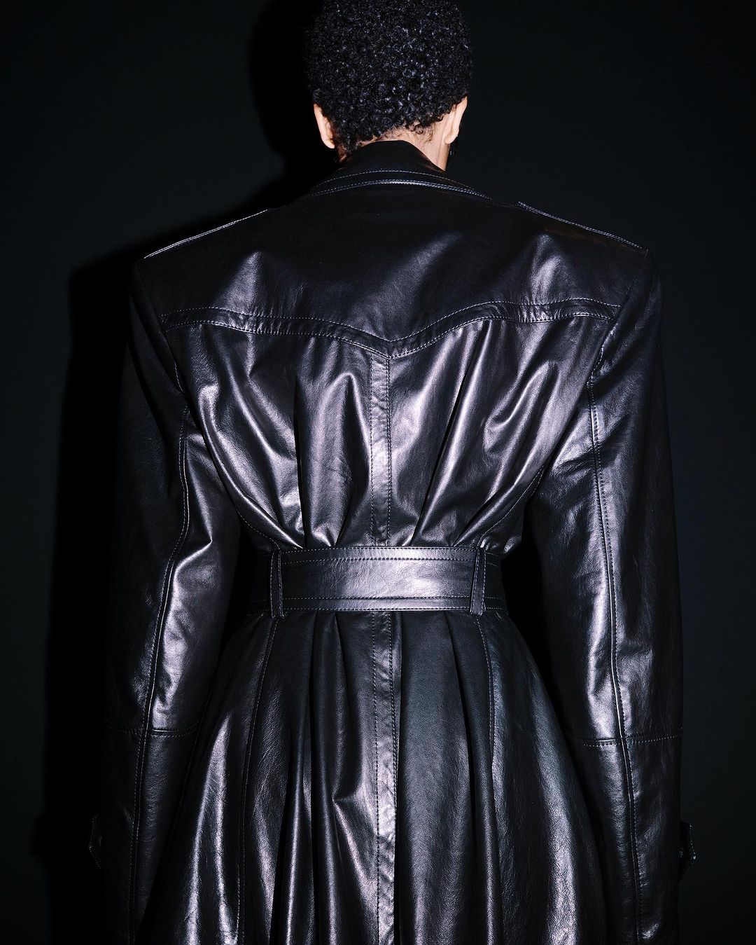 Back view of an adult wearing a black, belted, shiny leather trench coat, captured indoors against a dark background.