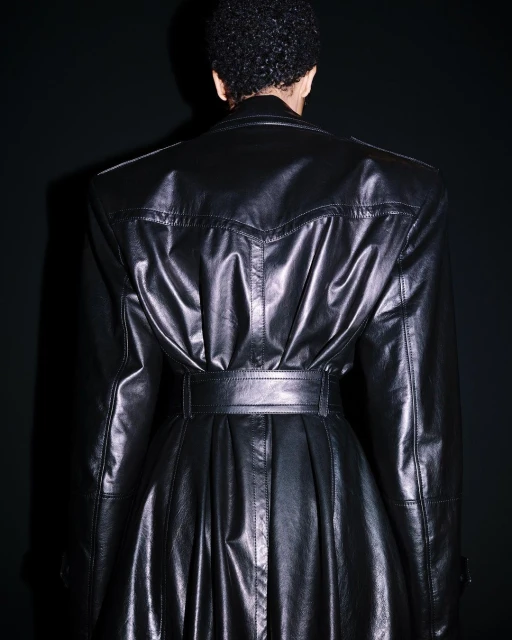 Back view of an adult wearing a black, belted, shiny leather trench coat, captured indoors against a dark background.