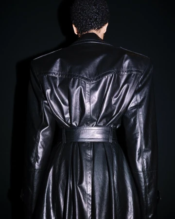 Back view of an adult wearing a black, belted, shiny leather trench coat, captured indoors against a dark background.