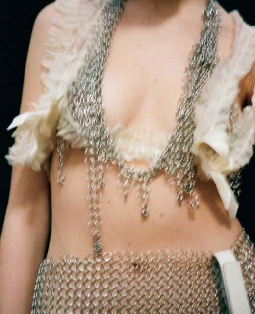 Closeup of a model in a chainmail top and skirt with ruffled details, set against a dark backdrop, emphasizing fashion and texture.