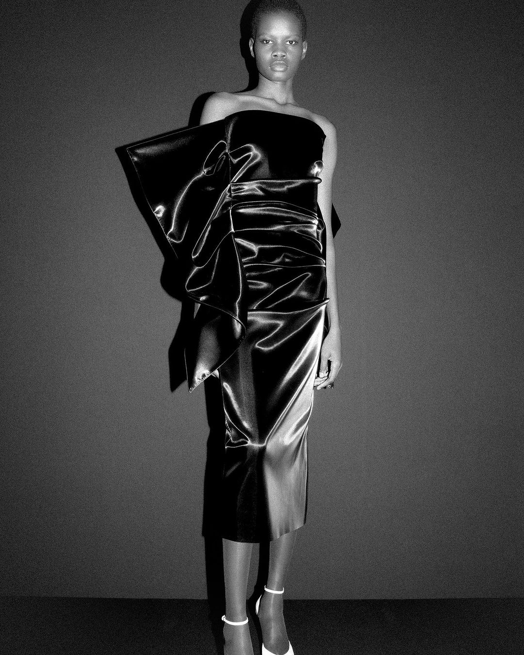 Black and white shot of an African female model wearing a black satin dress with a large bow and white high heels in a studio.