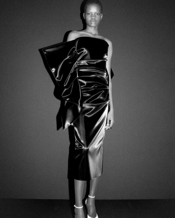 Black and white shot of an African female model wearing a black satin dress with a large bow and white high heels in a studio.