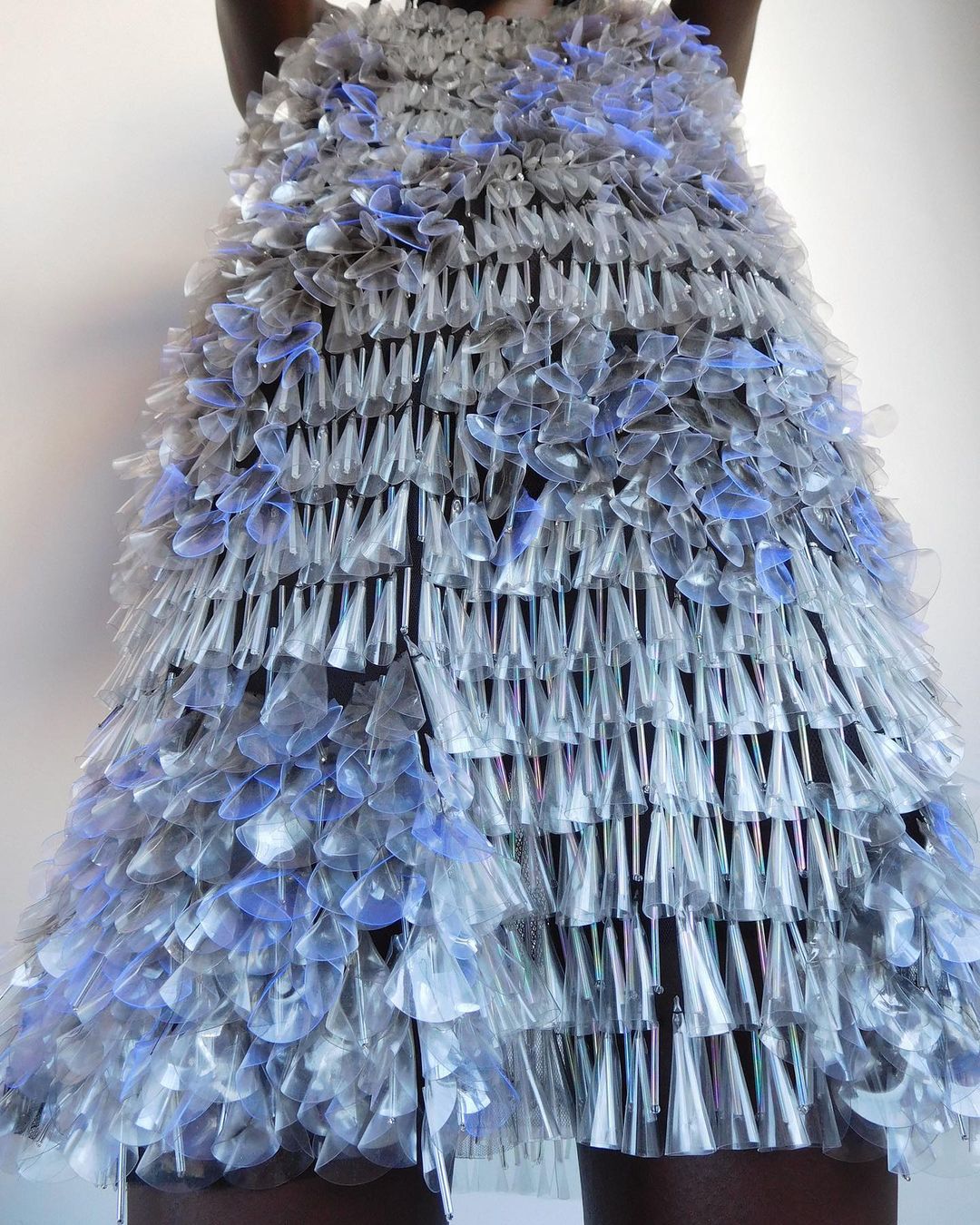 A woman poses in a dress made of clear and light blue plastic cups in a studio setting, showcasing avant-garde sustainable fashion.