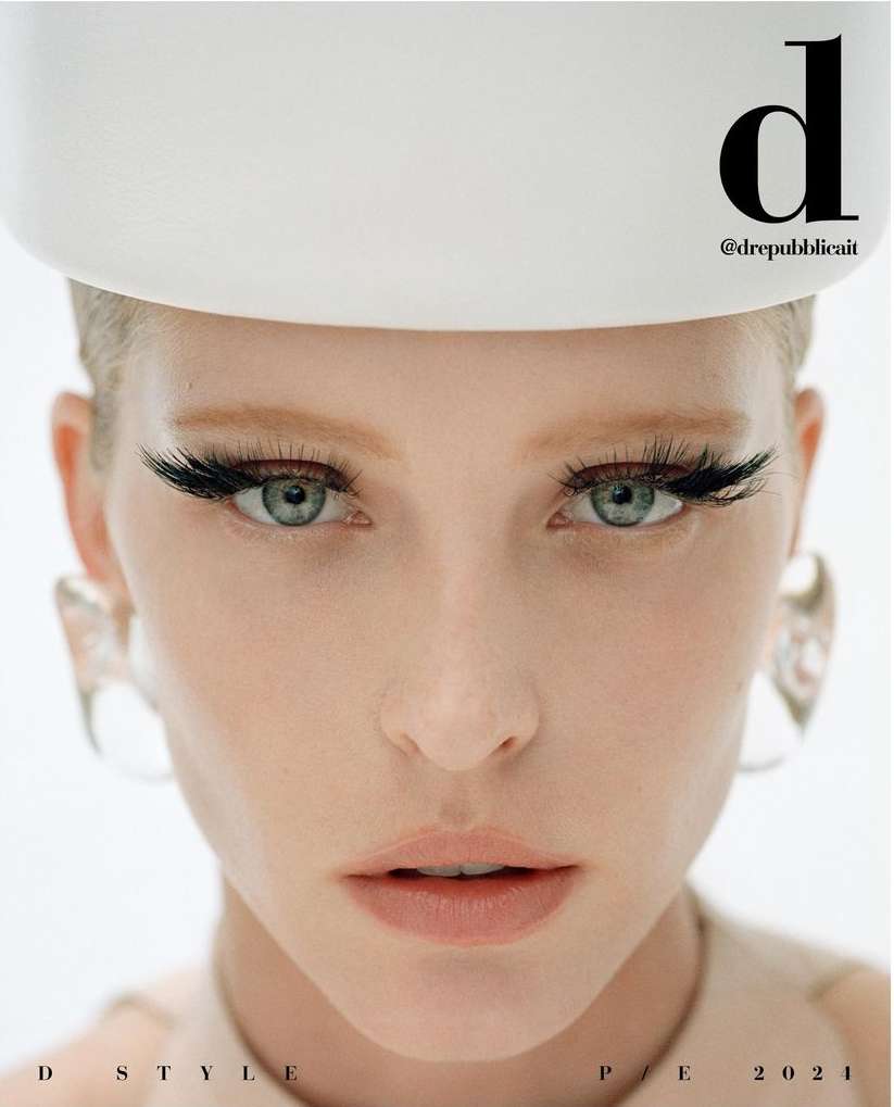 Close-up studio shot of a fair-skinned woman wearing a white hat and large false eyelashes, along with clear sculptural earrings. 
