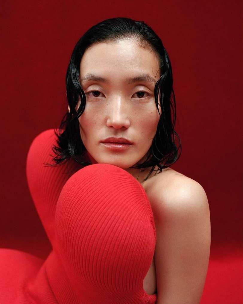 Close-up portrait of a woman with dark wavy hair and light skin, posed in front of a red background, wearing a red sweater.