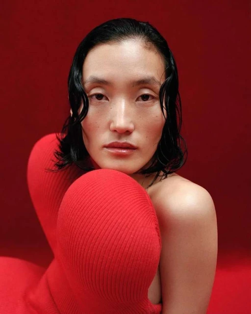 Close-up portrait of a woman with dark wavy hair and light skin, posed in front of a red background, wearing a red sweater.