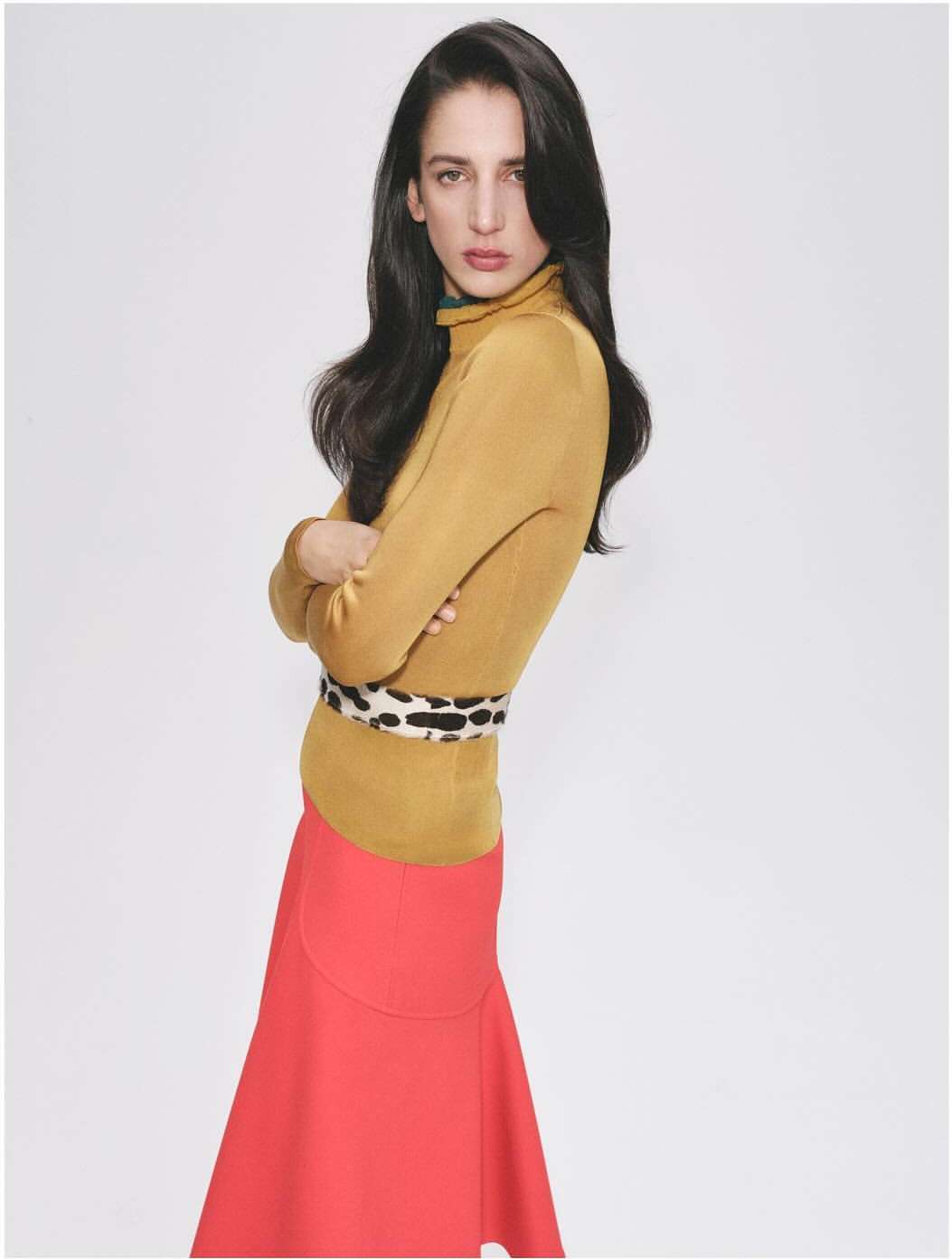 A young, light-skinned adult female model poses in a studio wearing a mustard yellow turtleneck sweater, a red midi skirt, and a patterned belt.