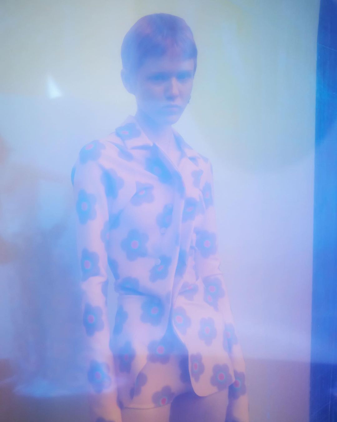 Portrait of a young adult female with short hair in a floral print jacket against a hazy, softly lit blue and yellow background.