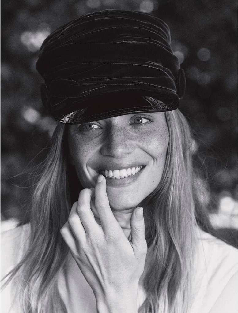 Black and white portrait of a smiling woman with blonde hair wearing a black hat, with her fingers touching her lips.
